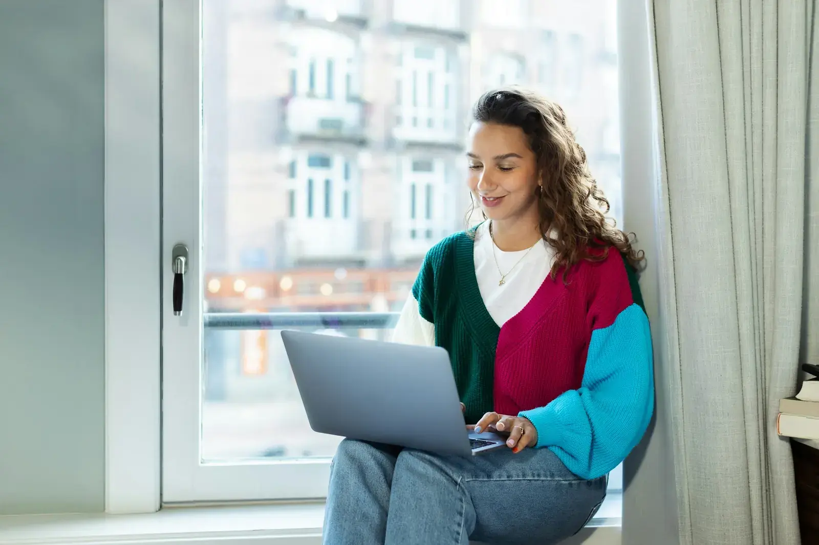 Una giovane donna seduta sul davanzale di una finestra lavora al computer portatile, sorridendo.